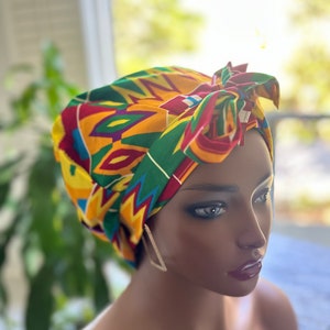 Op de afbeelding: Een kleurrijke hoofddoek met een levendig geometrisch patroon in rood, geel, groen en paars. De stof is bovenop het hoofd in een knoop geknoopt. De hoofddoek is een modeaccessoire.