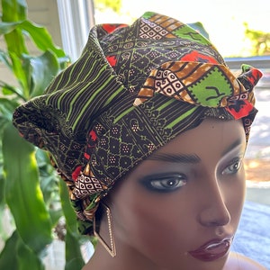 Op de afbeelding: Een patroon hoofddoek in zwart, groen, oranje en bruin. De stof heeft geometrische en gestreepte patronen. De hoofddoek wordt gedragen op een mannequinhoofd, gecombineerd met ruitvormige oorbellen.