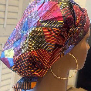 Op de afbeelding: Een kleurrijke hoofddoek met een geometrisch patroon in rood, blauw, geel en oranje. De hoofddoek is aan de achterkant geknoopt en wordt op een mannequinhoofd gedragen. Grote gouden oorringen zijn zichtbaar.