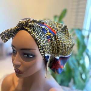 Op de afbeelding: Een hoofddoek met een geometrisch patroon in geel, zwart, rood en blauw. De stof heeft een geometrisch ontwerp en is in een strik geknoopt. De hoofddoek wordt op een mannequin gedragen.