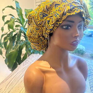 Op de afbeelding: Een patroonmuts in goud, blauw en wit, weergegeven op een mannequin. De muts heeft een ingewikkeld ontwerp. De mannequin staat op een houten oppervlak, met een plant op de achtergrond.