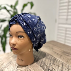 Op de afbeelding: Een marineblauwe tulband met een wit en lichtblauw cirkelpatroon wordt getoond op een mannequinhoofd. De tulband is gewikkeld en geknoopt, waardoor een stijlvolle hoofdbedekking ontstaat. Gouden oorringen worden door de mannequin gedragen.