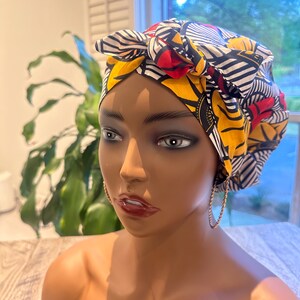 Op de afbeelding: Een hoofddoek met een levendig patroon van gele, rode, zwarte en witte geometrische vormen. De hoofddoek is bovenaan met een strik vastgebonden. De mannequin draagt gouden oorringen.