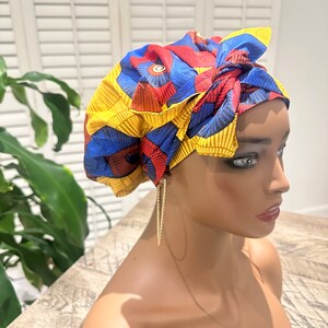 Op de afbeelding: Een kleurrijke hoofddoek met een geometrisch patroon in rood, blauw en geel. De stof is bovenop het hoofd geknoopt. De hoofddoek staat op een mannequinhoofd. Gouden oorringen zijn zichtbaar.