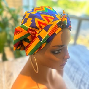 Op de afbeelding: Een kleurrijke hoofddoek met een geometrisch patroon in geel, blauw, rood en groen. De hoofddoek is bovenop geknoopt. De hoofddoek wordt gedragen door een mannequin.