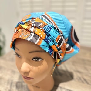 Op de afbeelding: Een patroon hoofddoek in blauw, oranje en geel. De stof heeft geometrische patronen en is gestyled op een mannequinhoofd. Deze levendige hoofddoek is een stijlvol accessoire.