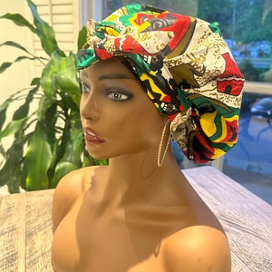 Op de afbeelding: Een kleurrijke bonnet met een levendige Afrikaanse print met rode, groene, gele en zwarte patronen. De bonnet wordt gedragen door een mannequin met gouden oorringen. De achtergrond omvat een raam en groen.