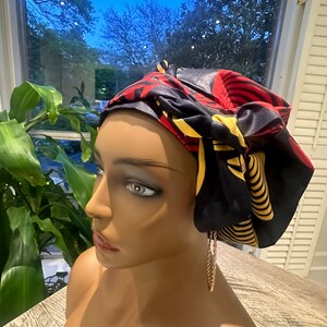 Op de afbeelding: Een zwart, rood en geel patroon hoofddoek is gebonden op een mannequin hoofd. De hoofddoek heeft een satijnen afwerking. Goudkleurige oorbellen hangen aan het oor van de mannequin. De achtergrond omvat een raam en groen.