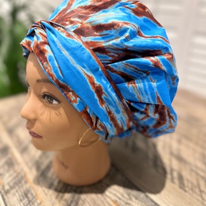 Op de afbeelding: Een blauw en bruin patroon hoofddoek. De hoofddoek is gemaakt van een zijdeachtig materiaal en is in tulbandstijl geknoopt. De hoofddoek zit op een mannequinhoofd met een neutrale huidskleur.