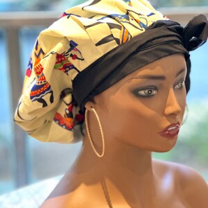 Op de afbeelding: Een crèmekleurige muts met een zwarte band en strik. De muts heeft een kleurrijk patroon van figuren. Het model draagt gouden oorringen. De achtergrond is wazig.