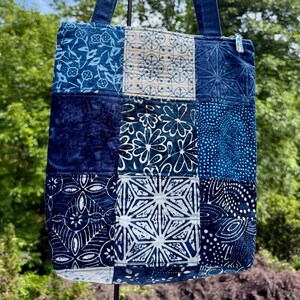 May include: A patchwork tote bag with a variety of blue fabric squares. The squares feature floral, geometric, and abstract patterns. The bag has denim handles and a zipper closure. The background shows a blue sky and green trees.