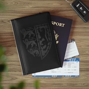 May include: Black passport holder featuring a shield design with three lions. A blue passport, two boarding passes, and a car key rest on a wooden surface. The passport holder has a textured finish.