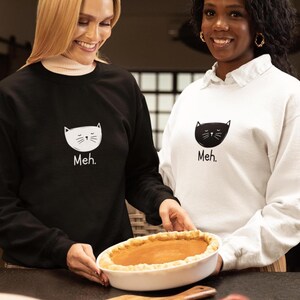 May include: A black sweatshirt with a white cat face graphic and the text "Meh." and a white sweatshirt with a black cat face graphic and the text "Meh." Both garments feature long sleeves, crew necklines, and a simple, clean design.