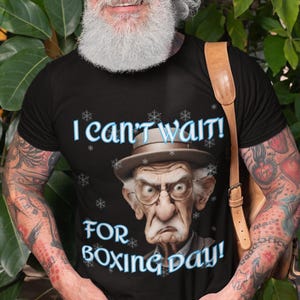 Könnte beinhalten: Schwarzes T-Shirt mit dem Text "I can't wait! FOR BOXING DAY!" und einer Karikatur eines alten Mannes mit Hut und Brille. Das T-Shirt wird von einer Person mit Tätowierungen und Bart getragen.