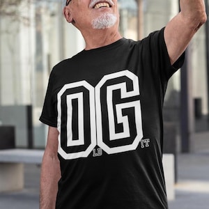以下が含まれることがあります： 黒の半袖Tシャツで、大きな白いブロック体の文字でOGと書かれています。OとGの文字の下部には小さなLDとITの文字が配置されており、グラフィックデザインでOLD SCHOOLという言葉を形成しています。