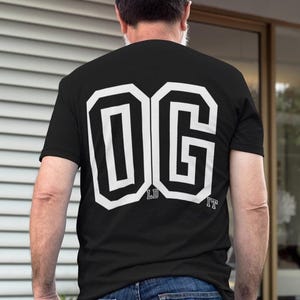 Op de afbeelding: Een zwart T-shirt met korte mouwen en grote witte blokletters op de achterkant die OG vormen. Kleinere witte tekst met de letters LD en IT staat onder de letters. Het T-shirt wordt gedragen met een blauwe spijkerbroek voor een casual look.