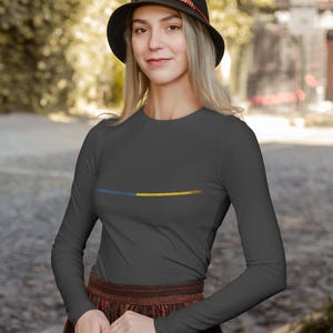 May include: A dark gray long-sleeve shirt with a horizontal blue and yellow design across the chest. The person is wearing a black hat with a brown band and a brown and red patterned skirt. The background is blurred.