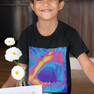 Op de afbeelding: Een jong persoon draagt een zwart T-shirt met een kleurrijke haai-afbeelding. De persoon houdt een kaart vast met de tekst "Happy Mother's Day" en er staan aardbeien en een bord met eieren op tafel.