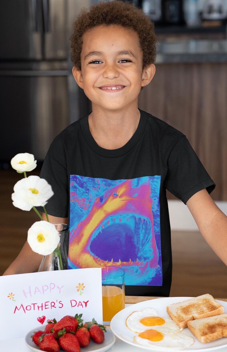 Op de afbeelding: Een jong persoon draagt een zwart T-shirt met een kleurrijke haai-afbeelding. De persoon houdt een kaart vast met de tekst "Happy Mother's Day" en er staan aardbeien en een bord met eieren op tafel.