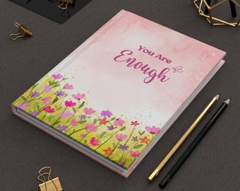 May include: A pink journal with a watercolor background and a floral border. The text "You Are Enough" is printed in pink on the front cover.