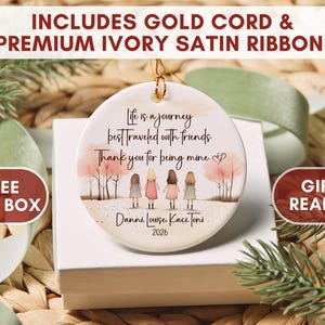 May include: A round white ceramic ornament featuring an illustration of four people standing in a row with pink trees in the background. The text reads: Life is a journey best traveled with friends. Thank you for being mine. Danni, Louise, Kaci, Toni, 2026.