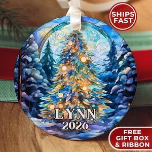 May include: A round ceramic ornament featuring a stained-glass style illustration of a glowing Christmas tree in a snowy forest. The text LYNN and 2026 is printed in black at the bottom. A white ribbon is attached to the top for hanging purposes.