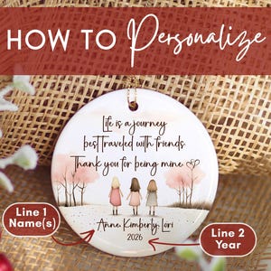 May include: A round white ceramic ornament featuring an illustration of three friends standing in a landscape with pink trees. The text reads: Life is a journey best traveled with friends. Thank you for being mine. Anne, Kimberly, Lori, and 2026.