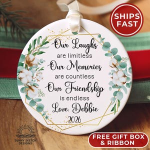 May include: A round white ceramic ornament featuring a gold geometric frame, green eucalyptus leaves, and white cotton flower illustrations. The text reads: Our Laughs are limitless, Our Memories are countless, Our Friendship is endless, Love, Debbie, 2026.