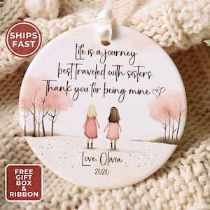May include: A round white ceramic ornament featuring a printed illustration of two figures standing in a landscape with pink trees. The text reads: Life is a journey best traveled with sisters. Thank you for being mine. Love, Olivia 2026.