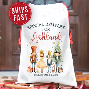 May include: White drawstring gift bag with red accents, featuring a Nutcracker-themed design. The bag is personalized with the name "Ashland" and the text "Special Delivery For" and "Love, Mommy & Daddy". A red circle reads "Ships Fast".