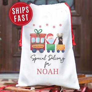 May include: White Christmas gift bag with a red drawstring closure. The bag features a colorful illustration of Santa Claus and a reindeer on a train, with the text "Special Delivery for NOAH". A red circle with "SHIPS FAST" is in the upper left.