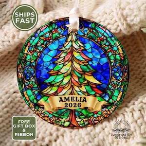 May include: A round, flat Christmas tree ornament featuring a colorful stained glass pattern with a central evergreen tree. A gold-colored ribbon banner across the bottom displays the name AMELIA and the year 2026 in black text. A white ribbon loop is attached.