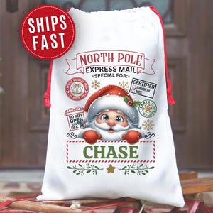 May include: White Christmas gift sack with a Santa Claus graphic. The sack features the text "North Pole Express Mail" and the name "Chase" in green. Red drawstring closure and accents.