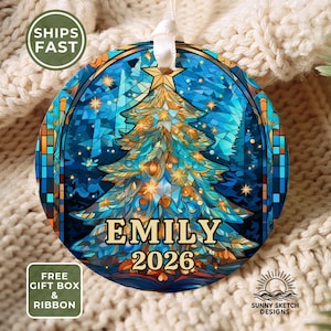 May include: A round ceramic ornament featuring a stained-glass style illustration of a Christmas tree in shades of blue and gold. The name EMILY and the year 2026 are printed in gold lettering at the bottom. A white ribbon is attached to the top.