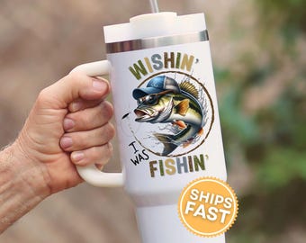 Może przedstawiać: Biały kubek termiczny z uchwytem, z kreskówkowym basem w okularach przeciwsłonecznych i kapeluszu. Tekst brzmi "Wishin' I Was Fishin'". Kubek ma białą pokrywkę i srebrny pasek.