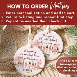May include: Two round white ceramic ornaments with white ribbon hangers. Each features a printed illustration of seven people holding hands, pink trees, and the text: Life is a journey best traveled with friends. Thank you for being mine. Love, Michelle 2026.