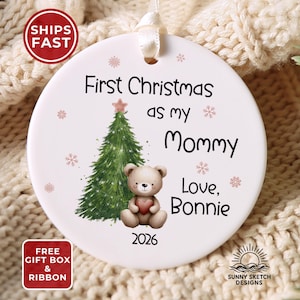May include: A round white ceramic ornament featuring a green Christmas tree with a pink star, a brown teddy bear holding a red heart, and the text First Christmas as my Mommy Love, Bonnie 2026, accented with small pink snowflakes.
