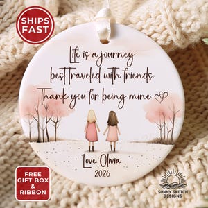 May include: A round white ceramic ornament featuring a printed illustration of two figures standing between pink trees. The text reads: Life is a journey best traveled with friends. Thank you for being mine. Love, Olivia 2026. It has a white ribbon.