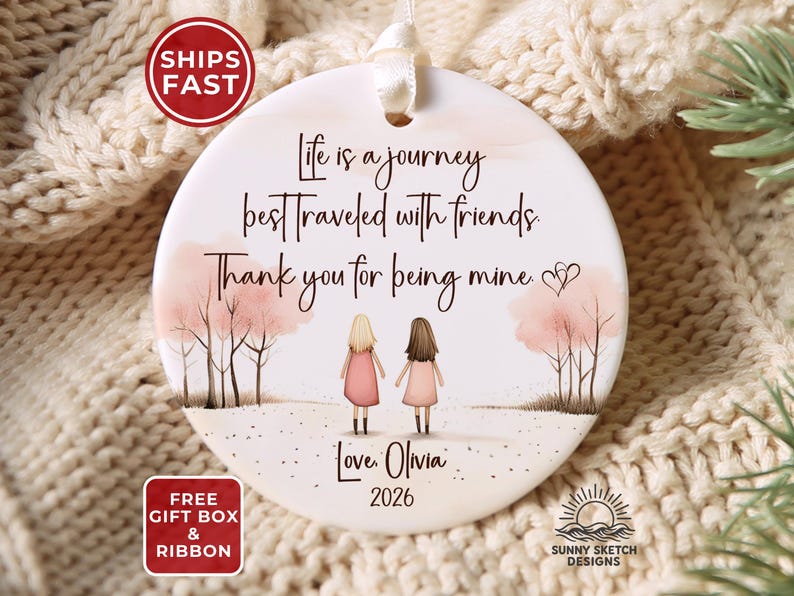 May include: A round white ceramic ornament featuring a printed illustration of two figures standing between pink trees. The text reads: Life is a journey best traveled with friends. Thank you for being mine. Love, Olivia 2026. It has a white ribbon.