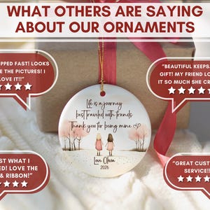 May include: A round white ceramic ornament featuring an illustration of two figures standing in a landscape with trees. The text reads: Life is a journey best traveled with friends. Thank you for being mine. Love, Olivia 2026. It hangs by a gold cord.