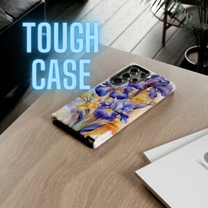 Può includere: Una custodia per telefono bianca con un design ad acquerello di iris viola e gialli. La custodia ha un rialzo per la fotocamera e il testo "TOUGH CASE" è visibile sullo sfondo.