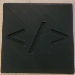 Könnte beinhalten: Ein schwarzes quadratisches Kunststoffstück mit einem erhabenen, schrägen, weißen, schrägen Schrägstrich und zwei weißen, schrägen Klammern, eine auf jeder Seite des Schrägstrichs.