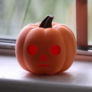 May include: A small orange pumpkin with a carved face and glowing red eyes. The pumpkin is sitting on a white surface.