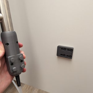 May include: A gray 3D printed tool with a silver metal rod and a white plastic tube attached to the bottom. The tool is held in a hand. A gray 3D printed wall mount with two holes is attached to a white wall.
