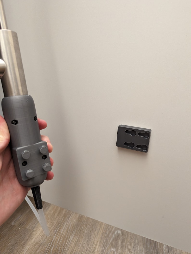 May include: A gray 3D printed tool with a silver metal rod and a white plastic tube attached to the bottom. The tool is held in a hand. A gray 3D printed wall mount with two holes is attached to a white wall.