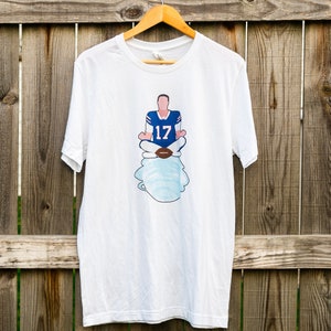 Puede incluir: Camiseta blanca con un gráfico de un jugador de fútbol americano con una camiseta azul con el número 17 sentado en posición de loto sobre una nube.