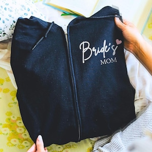 May include: A black zip-up hoodie featuring a white script and block letter graphic that reads "Bride's MOM" with a small pink heart icon. The garment includes a central metal zipper and black drawstring cords at the hood neckline.