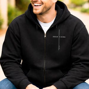 May include: Black zip-up hoodie featuring the text "JESUS IS KING" and a vertical line. The hoodie has a white inner layer and a metal zipper. The design is printed on the front of the hoodie.