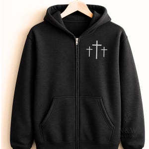 May include: Black zip-up hoodie with a hood and front pockets. The hoodie features a white graphic of three crosses on the upper left chest. The hoodie is hanging on a wooden hanger.