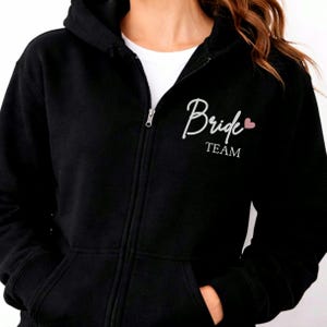 May include: A black zip-up hoodie featuring a white script font that reads Bride and the word TEAM in a block font underneath. A small light pink heart is positioned to the right of the word Bride on the upper left chest area of the garment.